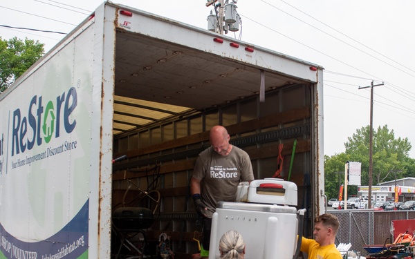 Three workers are loading or unloading a large white box truck belonging to Habitat for Humanity's ReStore program, a home improvement discount store. They are handling what appears to be a vintage or older-model washing machine at the rear of the truck. The scene takes place in a parking or staging area surrounded by various donated items and equipment.