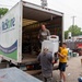 Three workers are loading or unloading a large white box truck belonging to Habitat for Humanity's ReStore program, a home improvement discount store. They are handling what appears to be a vintage or older-model washing machine at the rear of the truck. The scene takes place in a parking or staging area surrounded by various donated items and equipment.