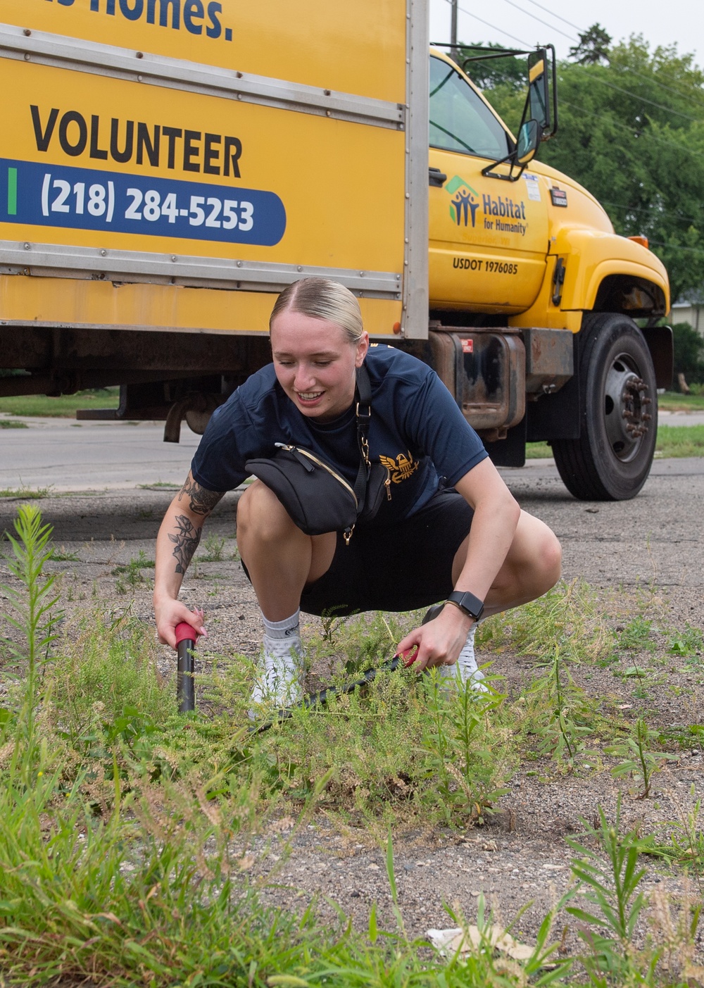 A young woman with blonde hair is crouching down and using hand tools to remove weeds or perform ground-level landscaping work. She is smiling and appears to be volunteering, as evidenced by the yellow Habitat for Humanity truck visible in the background. The setting appears to be a parking lot or roadside area with overgrown weeds.