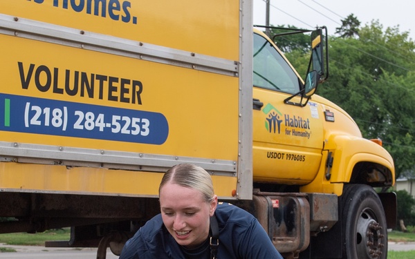 A young woman with blonde hair is crouching down and using hand tools to remove weeds or perform ground-level landscaping work. She is smiling and appears to be volunteering, as evidenced by the yellow Habitat for Humanity truck visible in the background. The setting appears to be a parking lot or roadside area with overgrown weeds.