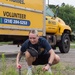A young woman with blonde hair is crouching down and using hand tools to remove weeds or perform ground-level landscaping work. She is smiling and appears to be volunteering, as evidenced by the yellow Habitat for Humanity truck visible in the background. The setting appears to be a parking lot or roadside area with overgrown weeds.