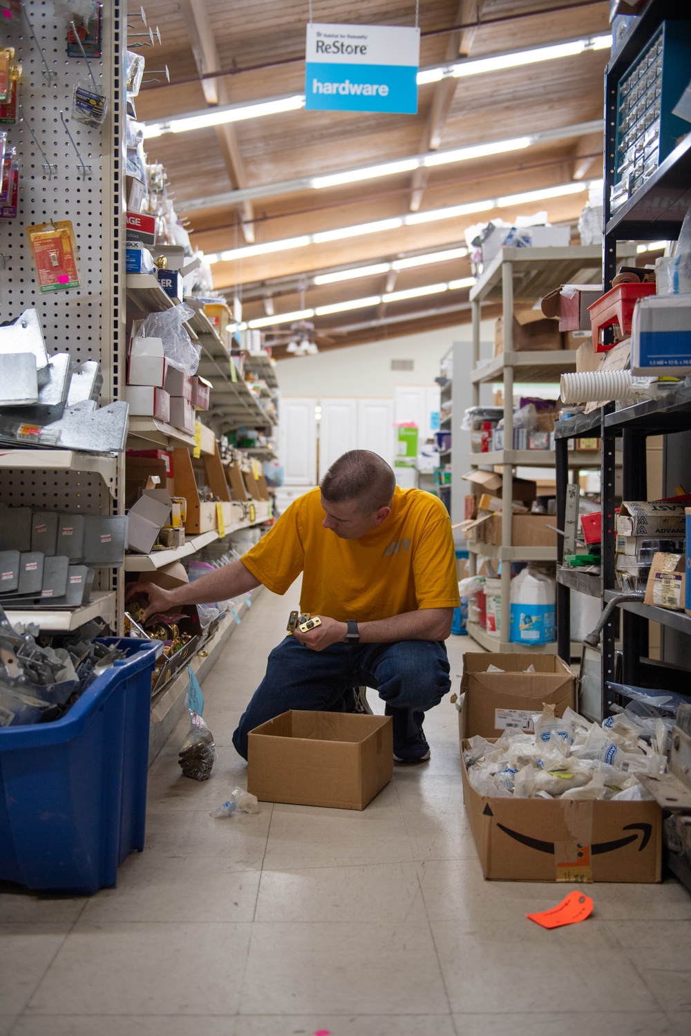 A man in a yellow t-shirt is crouching in the hardware aisle of a Habitat for Humanity ReStore, examining a small metal item, likely a hinge or latch. He is surrounded by shelves stocked with various hardware items, bins, and cardboard boxes including an Amazon box. The store appears to be a resale/thrift outlet for building materials and hardware.