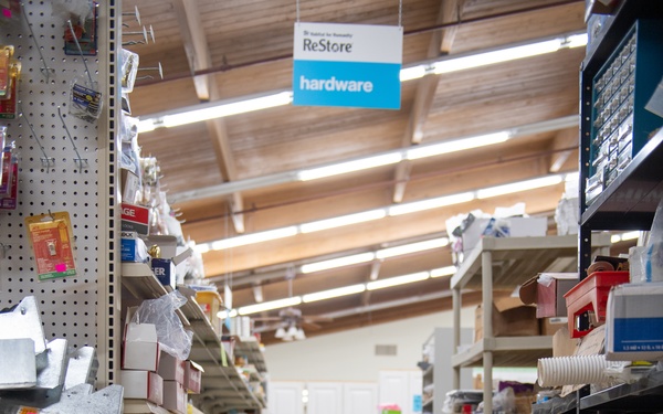 A man in a yellow t-shirt is crouching in the hardware aisle of a Habitat for Humanity ReStore, examining a small metal item, likely a hinge or latch. He is surrounded by shelves stocked with various hardware items, bins, and cardboard boxes including an Amazon box. The store appears to be a resale/thrift outlet for building materials and hardware.
