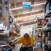 A man in a yellow t-shirt is crouching in the hardware aisle of a Habitat for Humanity ReStore, examining a small metal item, likely a hinge or latch. He is surrounded by shelves stocked with various hardware items, bins, and cardboard boxes including an Amazon box. The store appears to be a resale/thrift outlet for building materials and hardware.