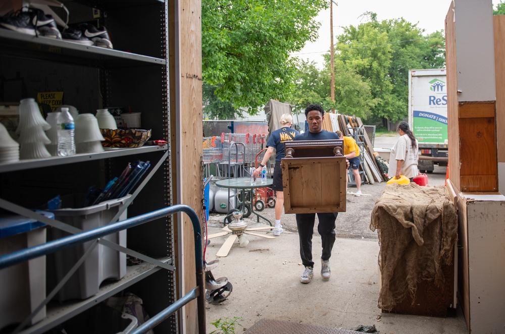 The image shows volunteers working at what appears to be a Habitat for Humanity ReStore donation center. A young man wearing a Navy t-shirt is carrying a wooden cabinet or nightstand into the building, while other volunteers are visible in the background handling various donated items. The outdoor area is filled with assorted furniture, building materials, and household goods.