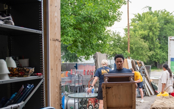 The image shows volunteers working at what appears to be a Habitat for Humanity ReStore donation center. A young man wearing a Navy t-shirt is carrying a wooden cabinet or nightstand into the building, while other volunteers are visible in the background handling various donated items. The outdoor area is filled with assorted furniture, building materials, and household goods.