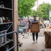 The image shows volunteers working at what appears to be a Habitat for Humanity ReStore donation center. A young man wearing a Navy t-shirt is carrying a wooden cabinet or nightstand into the building, while other volunteers are visible in the background handling various donated items. The outdoor area is filled with assorted furniture, building materials, and household goods.