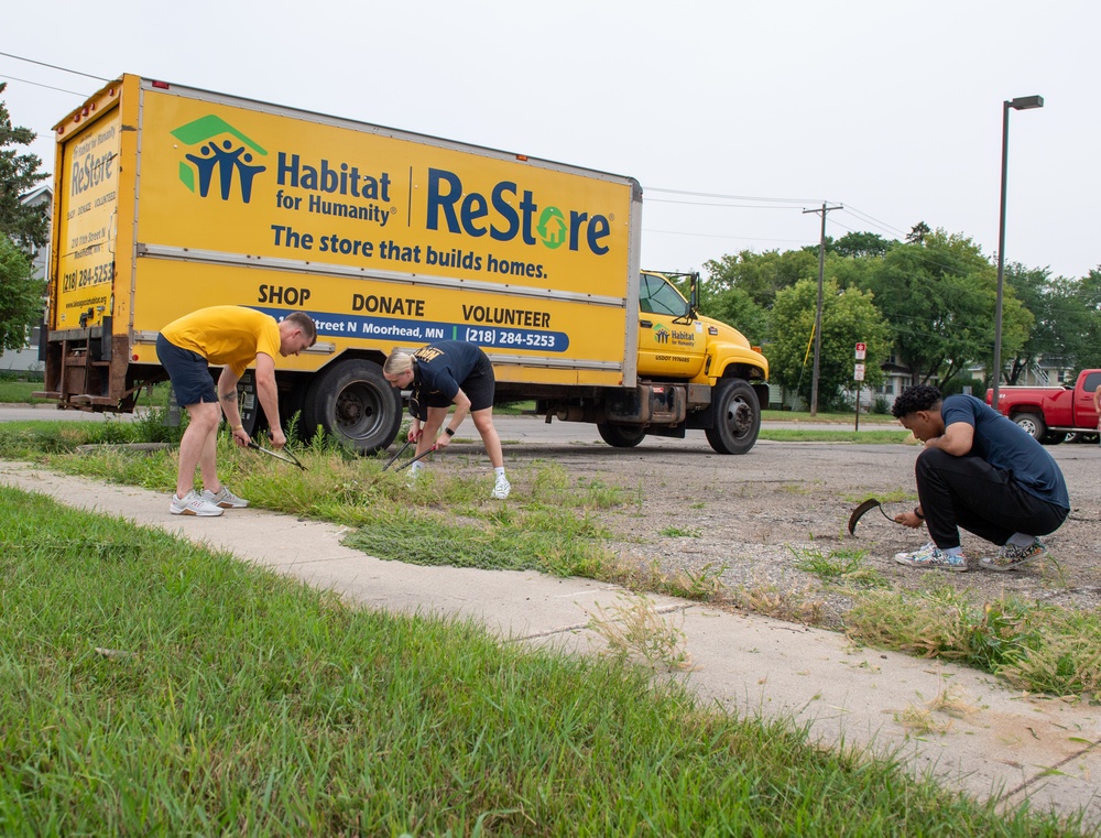 Three volunteers are performing outdoor landscaping or weeding work along a sidewalk in front of a large yellow Habitat for Humanity ReStore box truck. The truck is branded with the ReStore logo and displays the address in Moorhead, MN with phone number 218-284-5253. The scene appears to be a community service or volunteer cleanup event.