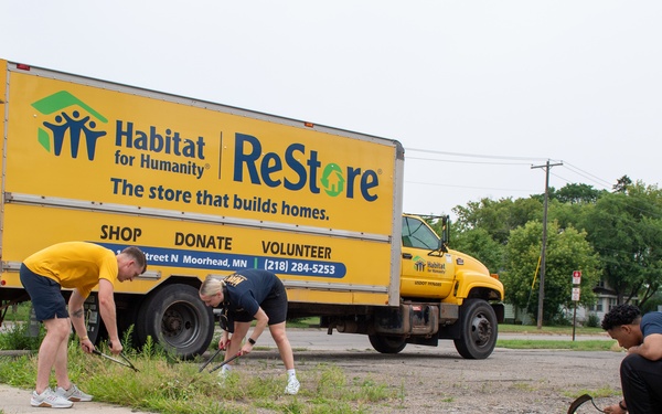 Three volunteers are performing outdoor landscaping or weeding work along a sidewalk in front of a large yellow Habitat for Humanity ReStore box truck. The truck is branded with the ReStore logo and displays the address in Moorhead, MN with phone number 218-284-5253. The scene appears to be a community service or volunteer cleanup event.