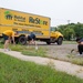 Three volunteers are performing outdoor landscaping or weeding work along a sidewalk in front of a large yellow Habitat for Humanity ReStore box truck. The truck is branded with the ReStore logo and displays the address in Moorhead, MN with phone number 218-284-5253. The scene appears to be a community service or volunteer cleanup event.