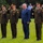 A formal military ceremony taking place outdoors on a grass lawn, featuring three uniformed military officers and one civilian in a suit standing in a line. The military personnel are rendering salutes simultaneously, suggesting a ceremonial event such as a wreath-laying, national commemoration, or official review. Civilian onlookers, including children and adults, are visible in the background.