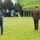 An outdoor military ceremony taking place on a grass lawn, likely a formal presentation or award ceremony. A civilian in a blue suit is speaking at a microphone podium while holding a document, facing two senior military officers in olive/khaki dress uniforms. A group of military personnel and civilians observe in the background.