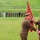 A military ceremony is taking place outdoors on a grassy field, where two senior officers in olive/khaki uniforms are conducting what appears to be a flag or colors transfer ceremony. A formation of military and civilian officials stands in the background observing the proceedings. A stone statue or monument is visible in the upper right background, and an iron fence lines the perimeter.