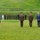 A formal military ceremony is taking place outdoors on a grass field, with several senior military officers in olive/khaki dress uniforms standing at attention. A officer at a microphone stand appears to be delivering a speech, while two senior officers and a civilian in a blue suit stand nearby. A group of approximately 20 military personnel from various branches and nations are assembled in the background near a metal fence, with a bronze statue visible on the right.