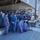 A group of military personnel in blue digital camouflage uniforms, along with individuals in civilian and multicam/tactical clothing, are gathered on the deck of a naval vessel. Most participants are wearing face masks, suggesting this was taken during the COVID-19 pandemic period. The group appears to be engaged in a briefing, demonstration, or training exercise around equipment cases on the ship's deck.