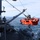A bright orange submersible or underwater vehicle decorated with a shark mouth design is being deployed from a naval or research vessel using a crane/davit system. The vehicle is suspended over open ocean water alongside the ship's hull. Two crew members in safety gear are visible on the ship's deck overseeing the operation.