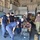 A group of military personnel from what appear to be multiple nations are gathered around tactical equipment cases in what looks like a ship's hangar or workshop bay. Several individuals are wearing face masks, suggesting this was taken during the COVID-19 pandemic period. The scene appears to depict a joint military training or equipment demonstration exercise.