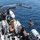 A naval vessel is conducting an at-sea operation involving the deployment or recovery of an orange buoy or spherical object using a deck crane. Several crew members in dark uniforms are visible on the deck overseeing the operation. The scene takes place in calm, open blue water under clear daylight conditions.