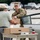 A U.S. Army soldier in OCP (Operational Camouflage Pattern) uniform is handing a cardboard box to a civilian man in a parking lot setting. A folding table in the foreground has cardboard boxes and is labeled 'BPC - Investigation' in handwritten text. The scene appears to be an outdoor distribution or evidence/materials handoff operation.