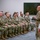 A military briefing or address is taking place in a modern conference room, where a senior officer in Army Combat Uniform (ACU/OCP pattern) stands and speaks to a large group of seated soldiers and some civilians. The audience is arranged in rows of chairs facing the speaker, who appears to be delivering remarks or a command briefing. The setting suggests a formal military gathering such as a unit briefing, promotion ceremony, or command visit.