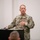 A U.S. Air Force officer in OCP (Operational Camouflage Pattern) uniform is delivering a briefing or speech at a podium to an audience of military personnel. The speaker's name tape is partially visible and the 'U.S. AIR FORCE' tape is clearly readable on the uniform. The setting appears to be an indoor briefing room or conference facility.