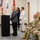 A formal official ceremony or briefing is taking place in what appears to be a government or military facility. A civilian woman in a gray blazer is speaking at a podium while two other civilian women stand behind her, and several U.S. Army soldiers in OCP (Operational Camouflage Pattern) uniforms are seated to the right. Multiple flags, including the U.S. flag and what appears to be a state flag and military organizational flags, are displayed in the background.