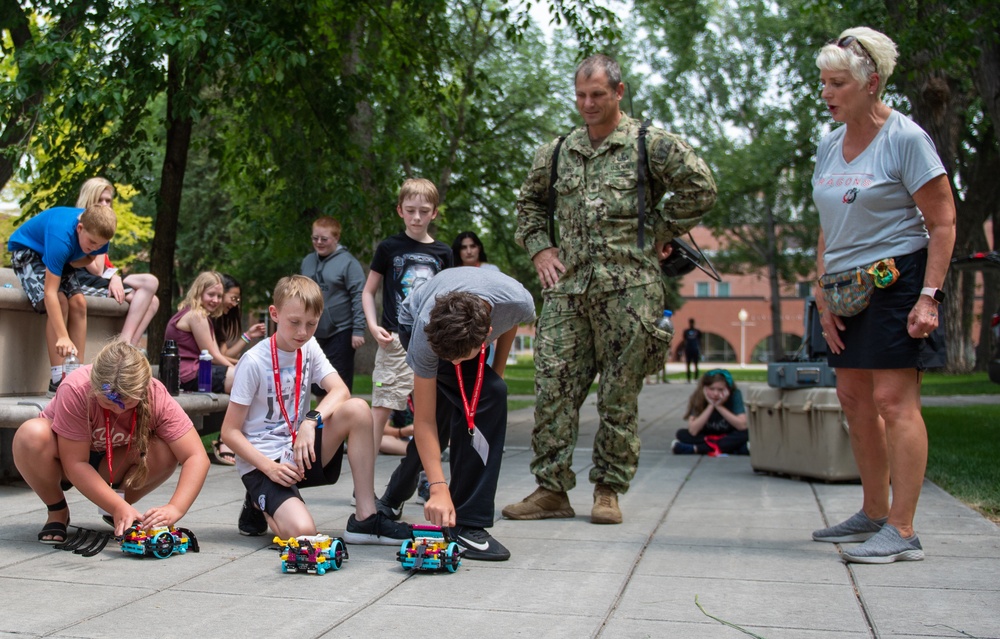 A group of children participate in an outdoor STEM or robotics activity on a campus or institutional grounds, placing small LEGO-style robotic vehicles on a paved walkway. A military personnel member in Navy Working Uniform (NWU) and a civilian woman supervisor observe the activity. The setting appears to be an educational event or camp, with multiple children wearing red lanyards.