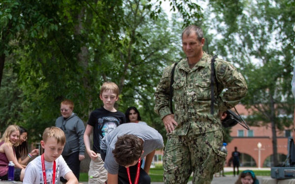 A group of children participate in an outdoor STEM or robotics activity on a campus or institutional grounds, placing small LEGO-style robotic vehicles on a paved walkway. A military personnel member in Navy Working Uniform (NWU) and a civilian woman supervisor observe the activity. The setting appears to be an educational event or camp, with multiple children wearing red lanyards.