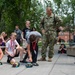 A group of children participate in an outdoor STEM or robotics activity on a campus or institutional grounds, placing small LEGO-style robotic vehicles on a paved walkway. A military personnel member in Navy Working Uniform (NWU) and a civilian woman supervisor observe the activity. The setting appears to be an educational event or camp, with multiple children wearing red lanyards.