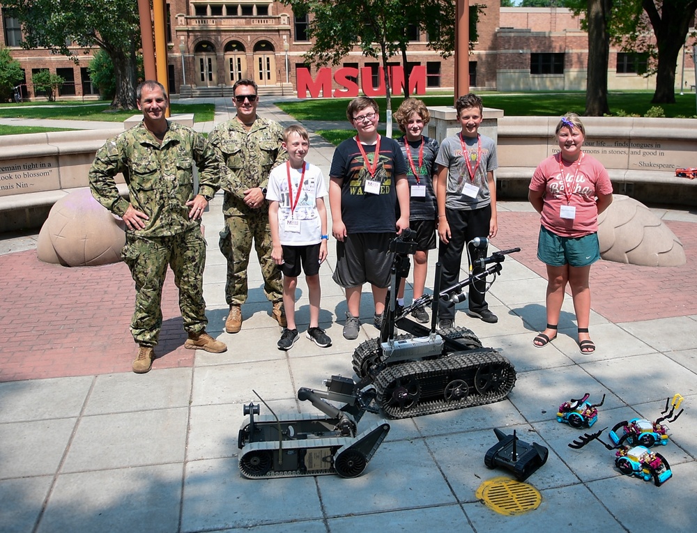 Two military personnel in camouflage uniforms pose with four children and one adult woman in front of an MSUM (Minnesota State University Moorhead) campus sign. The group is gathered around military-grade tracked robots, likely EOD (Explosive Ordnance Disposal) robots, along with smaller student-built robots on the ground. This appears to be a STEM or robotics educational event where military personnel are demonstrating equipment to young students.