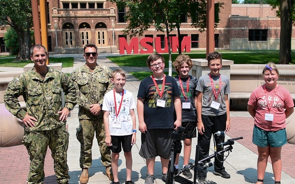 Two military personnel in camouflage uniforms pose with four children and one adult woman in front of an MSUM (Minnesota State University Moorhead) campus sign. The group is gathered around military-grade tracked robots, likely EOD (Explosive Ordnance Disposal) robots, along with smaller student-built robots on the ground. This appears to be a STEM or robotics educational event where military personnel are demonstrating equipment to young students.