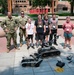 Two military personnel in camouflage uniforms pose with four children and one adult woman in front of an MSUM (Minnesota State University Moorhead) campus sign. The group is gathered around military-grade tracked robots, likely EOD (Explosive Ordnance Disposal) robots, along with smaller student-built robots on the ground. This appears to be a STEM or robotics educational event where military personnel are demonstrating equipment to young students.