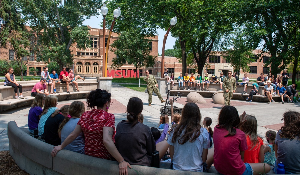 An outdoor demonstration is taking place on the campus of MSUM (Minnesota State University Moorhead), where two military personnel in camouflage uniforms are presenting what appears to be a small robotic device to a crowd of children and adults. The audience is seated on curved concrete benches in a campus plaza, attentively watching the presentation. The setting is a sunny day with mature trees and red brick university buildings visible in the background.