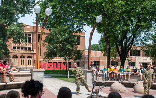 An outdoor demonstration is taking place on the campus of MSUM (Minnesota State University Moorhead), where two military personnel in camouflage uniforms are presenting what appears to be a small robotic device to a crowd of children and adults. The audience is seated on curved concrete benches in a campus plaza, attentively watching the presentation. The setting is a sunny day with mature trees and red brick university buildings visible in the background.