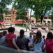 An outdoor demonstration is taking place on the campus of MSUM (Minnesota State University Moorhead), where two military personnel in camouflage uniforms are presenting what appears to be a small robotic device to a crowd of children and adults. The audience is seated on curved concrete benches in a campus plaza, attentively watching the presentation. The setting is a sunny day with mature trees and red brick university buildings visible in the background.