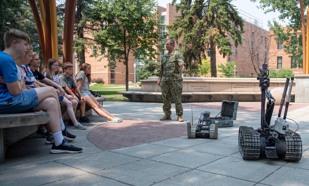 A military service member in Navy digital camouflage uniform is conducting an outdoor demonstration of robotic equipment to a group of young students seated on benches. Two robots are visible on the paved plaza area — one smaller tracked robot and one larger tracked robot with a mechanical arm. The setting appears to be a campus or institutional outdoor courtyard with brick buildings and trees in the background.