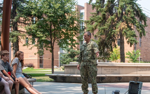 A military service member in Navy digital camouflage uniform is conducting an outdoor demonstration of robotic equipment to a group of young students seated on benches. Two robots are visible on the paved plaza area — one smaller tracked robot and one larger tracked robot with a mechanical arm. The setting appears to be a campus or institutional outdoor courtyard with brick buildings and trees in the background.