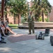 A military service member in Navy digital camouflage uniform is conducting an outdoor demonstration of robotic equipment to a group of young students seated on benches. Two robots are visible on the paved plaza area — one smaller tracked robot and one larger tracked robot with a mechanical arm. The setting appears to be a campus or institutional outdoor courtyard with brick buildings and trees in the background.