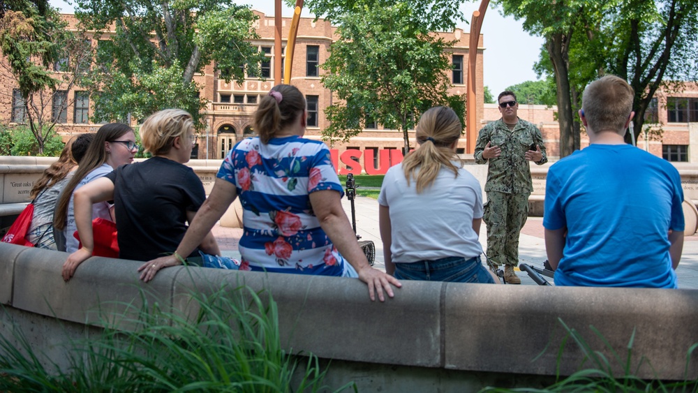 A person in U.S. Navy camouflage uniform is addressing a small group of civilians outdoors on what appears to be a university campus. The group of approximately five people is seated or leaning against a low concrete wall, listening to the uniformed individual speak. Large red letters in the background partially spell 'SU', suggesting this is likely a state university campus.