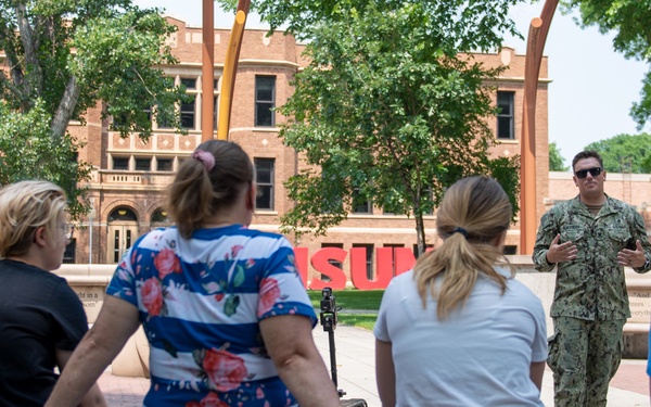 A person in U.S. Navy camouflage uniform is addressing a small group of civilians outdoors on what appears to be a university campus. The group of approximately five people is seated or leaning against a low concrete wall, listening to the uniformed individual speak. Large red letters in the background partially spell 'SU', suggesting this is likely a state university campus.