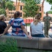 A person in U.S. Navy camouflage uniform is addressing a small group of civilians outdoors on what appears to be a university campus. The group of approximately five people is seated or leaning against a low concrete wall, listening to the uniformed individual speak. Large red letters in the background partially spell 'SU', suggesting this is likely a state university campus.