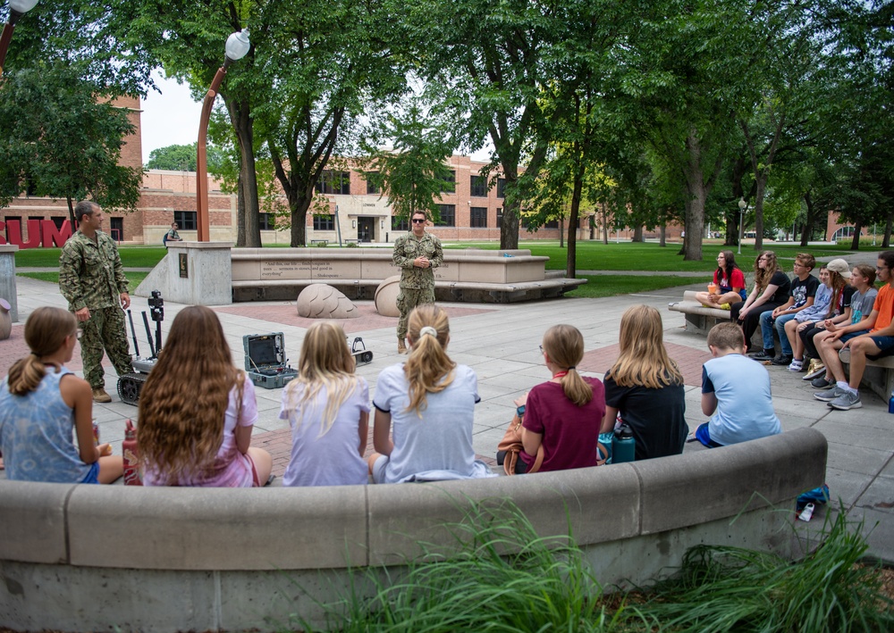 Two military personnel in camouflage uniforms are conducting an outdoor educational presentation for a group of children seated on curved concrete benches in a campus or park setting. A small ground robot and equipment cases are visible on the ground between the presenters and the audience. The setting appears to be a public institution, likely a university or military museum, based on the brick buildings and landscaped grounds visible in the background.