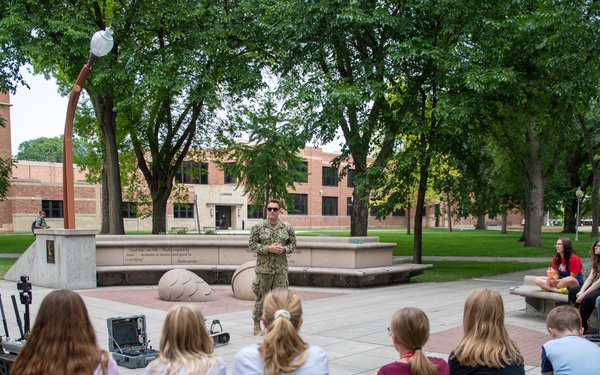 Two military personnel in camouflage uniforms are conducting an outdoor educational presentation for a group of children seated on curved concrete benches in a campus or park setting. A small ground robot and equipment cases are visible on the ground between the presenters and the audience. The setting appears to be a public institution, likely a university or military museum, based on the brick buildings and landscaped grounds visible in the background.