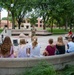 Two military personnel in camouflage uniforms are conducting an outdoor educational presentation for a group of children seated on curved concrete benches in a campus or park setting. A small ground robot and equipment cases are visible on the ground between the presenters and the audience. The setting appears to be a public institution, likely a university or military museum, based on the brick buildings and landscaped grounds visible in the background.