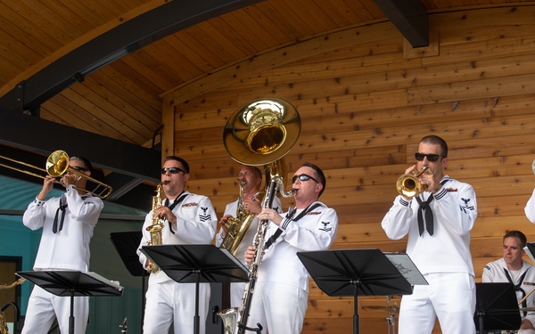 A military band composed of U.S. Navy sailors in white dress uniforms performs on an outdoor covered stage. The ensemble includes brass and woodwind instruments, with a drummer in the background. The performance appears to be taking place at a public venue identified as Broadway Square.