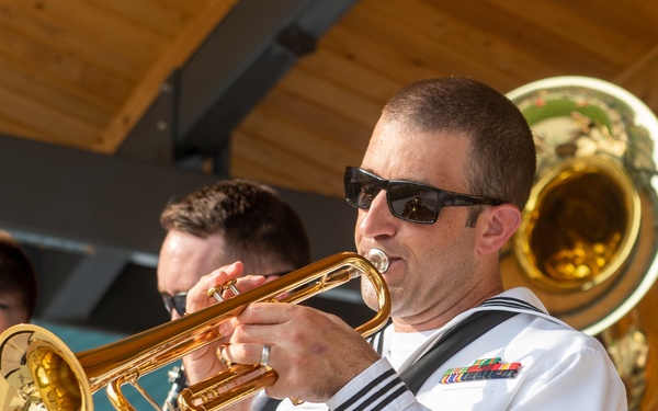 A group of U.S. Navy musicians in dress white uniforms are performing at an outdoor event. The foreground subject is a Petty Officer playing a gold trumpet, wearing sunglasses and displaying service ribbons and a rating badge. Additional band members are visible in the background playing clarinet and sousaphone.