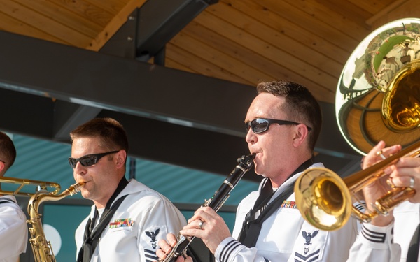 A group of U.S. Navy musicians in dress white uniforms are performing outdoors as a military band. Four musicians are visible in the foreground playing various instruments, with a fifth partially visible in the background. They are positioned in front of music stands at what appears to be an outdoor venue with wooden architectural elements.