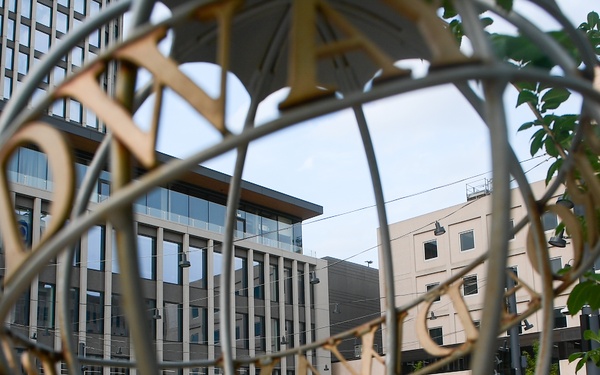 The image is taken through a large decorative metal globe sculpture with gold lettering, framing a view of a modern urban public plaza. In the background, a group of performers dressed in white uniforms are on a small wooden stage, with an audience seated in red chairs on the right. The scene includes a fountain, green lawn, trees, and surrounding multi-story commercial or office buildings.