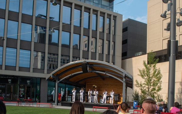 An outdoor public concert is taking place in an urban plaza, where a military band dressed in white uniforms performs on a covered bandshell stage. Audience members are seated at red bistro-style tables and chairs on a paved area adjacent to a green lawn. The setting appears to be a modern downtown district with commercial buildings, including a visible US Bank branch.