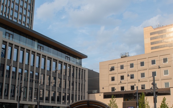 The image shows Broadway Square in Fargo, North Dakota, a modern urban public plaza featuring an interactive fountain and outdoor performance stage. People of various ages are gathered to watch what appears to be a live musical performance by a group dressed in white uniforms. The scene is set during golden hour with contemporary mixed-use buildings surrounding the square.